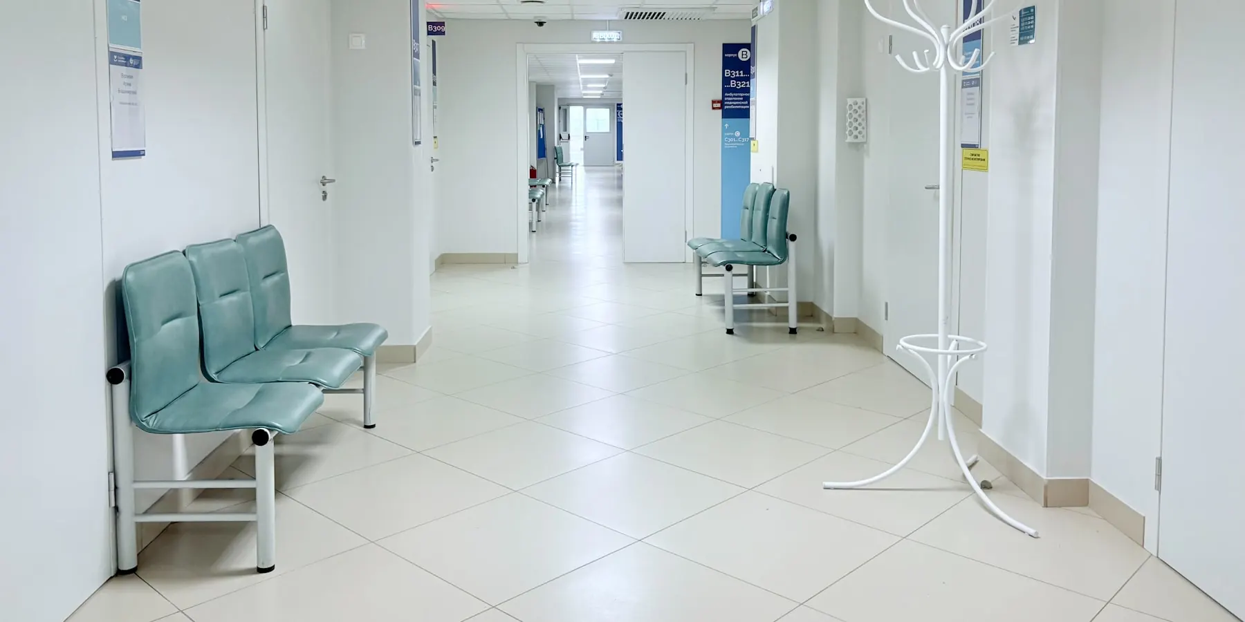 ทางเดินในโรงพยาบาลที่มีเก้าอี้นั่งรอเรียงอยู่ริมผนังและป้ายบอกหมายเลขห้องตรวจ